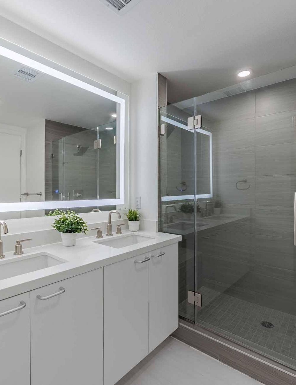 Evora modern bathroom with a double sink, large lit mirror, glass shower, and potted plants on the counter.