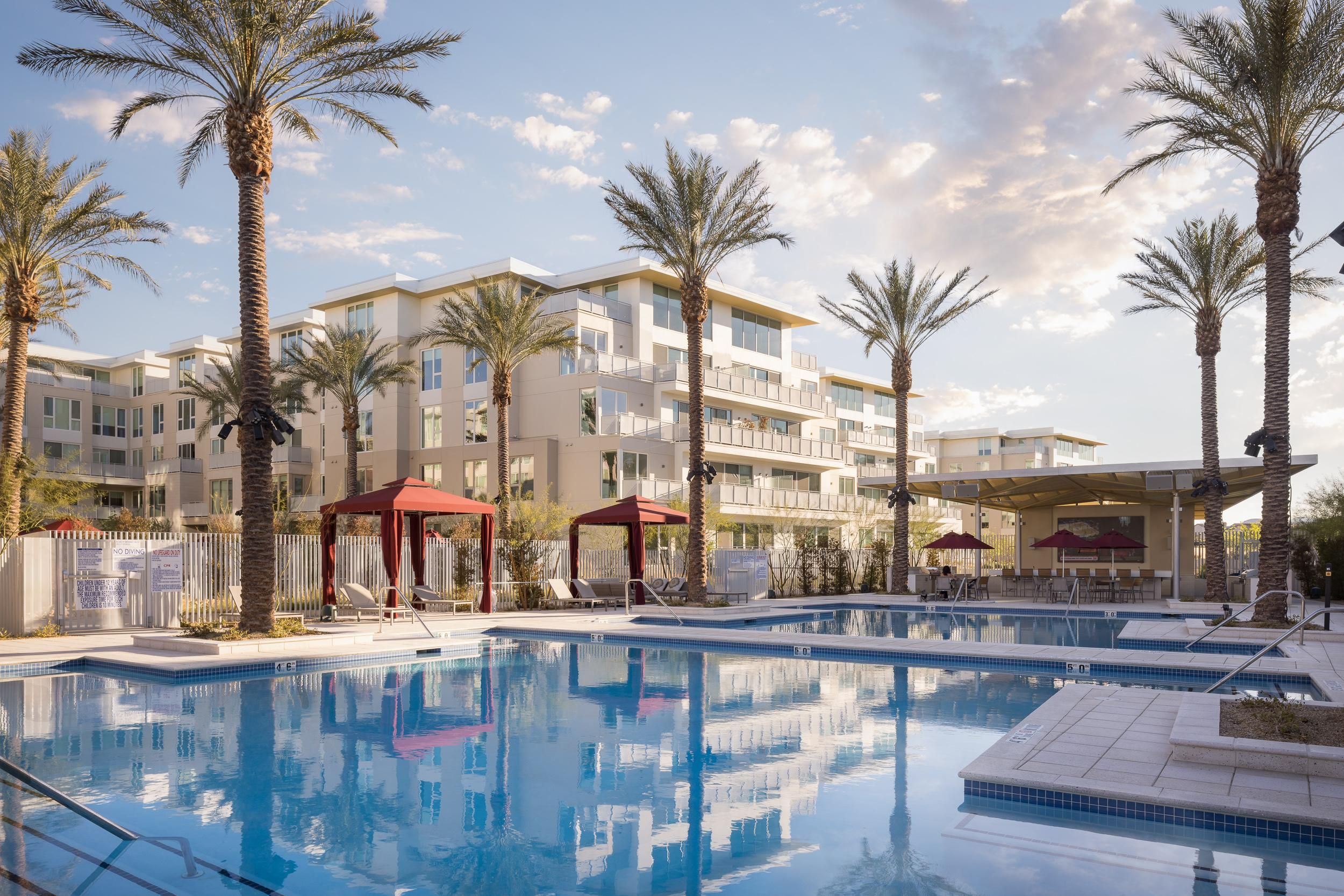 Luxury apartment buildings with palm trees surround a swimming pool with red cabanas and lounge chairs under a partly cloudy sky. Photo credit: Michael Tessler Photography.