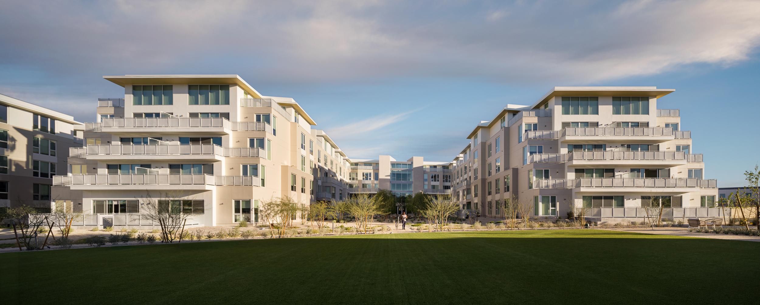Modern white apartment buildings with balconies surround a large green lawn under a partly cloudy sky. Photo credit: Michael Tessler Photography.