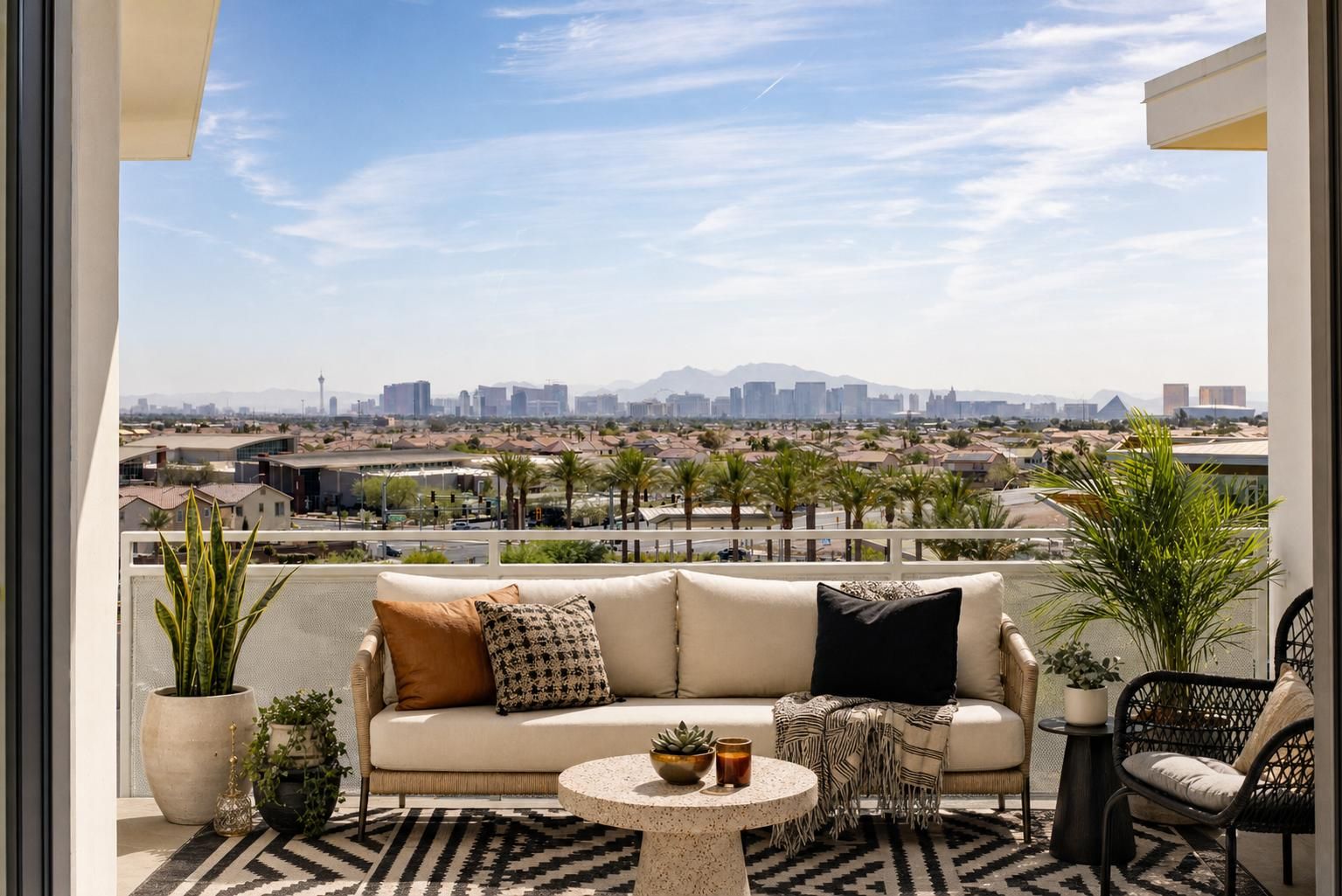 Outdoor patio with a sofa, plants, small table, and city skyline with mountains in the background under a blue sky.