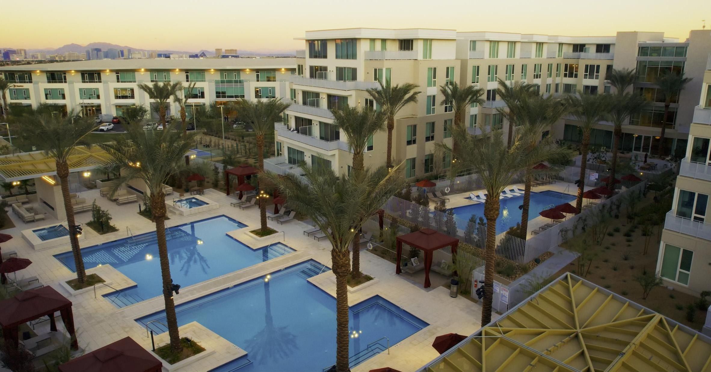 Modern apartment complex with multiple pools, palm trees, and lounge areas at sunset.