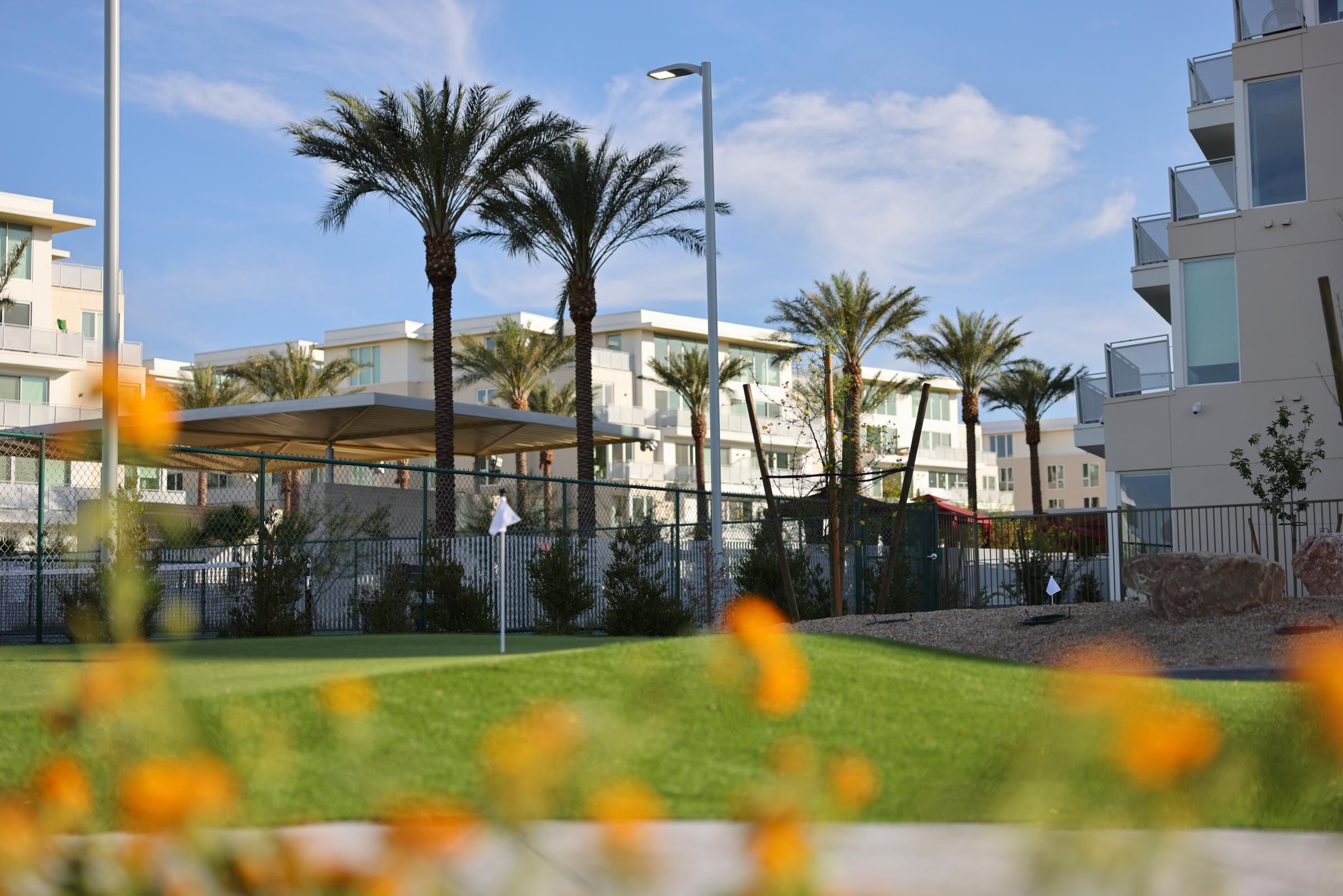 Modern apartment buildings with palm trees and green lawn; orange flowers blurred in the foreground.