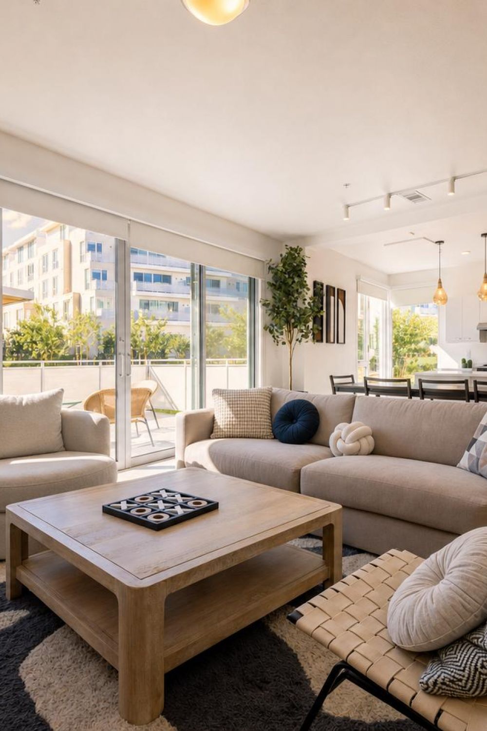 Bright modern living room with beige furniture, large windows, a dining area, and an open kitchen, overlooking a sunny outdoor patio.