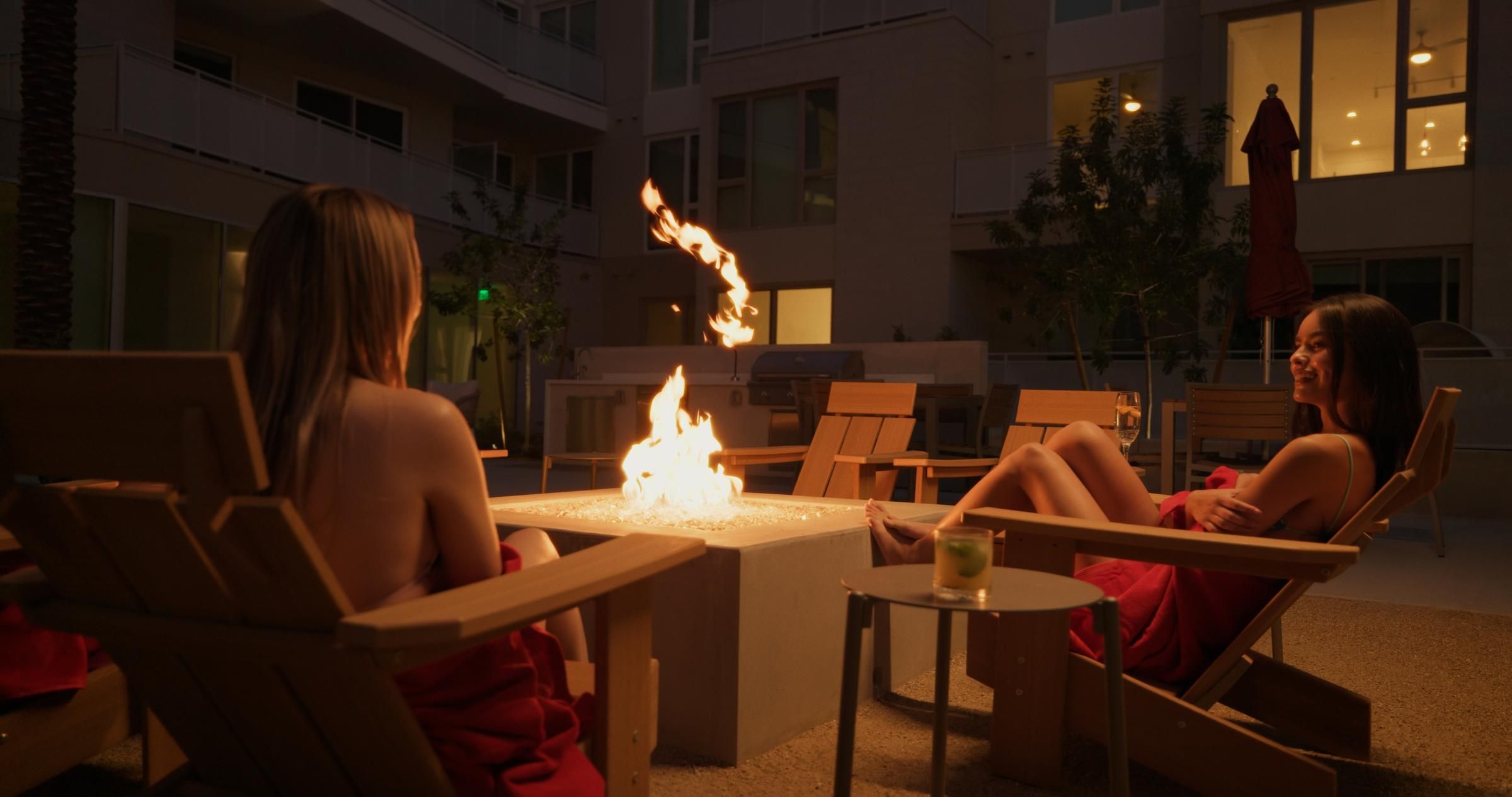 Two women in towels relax by an outdoor fire pit at night, sitting on chairs and talking.