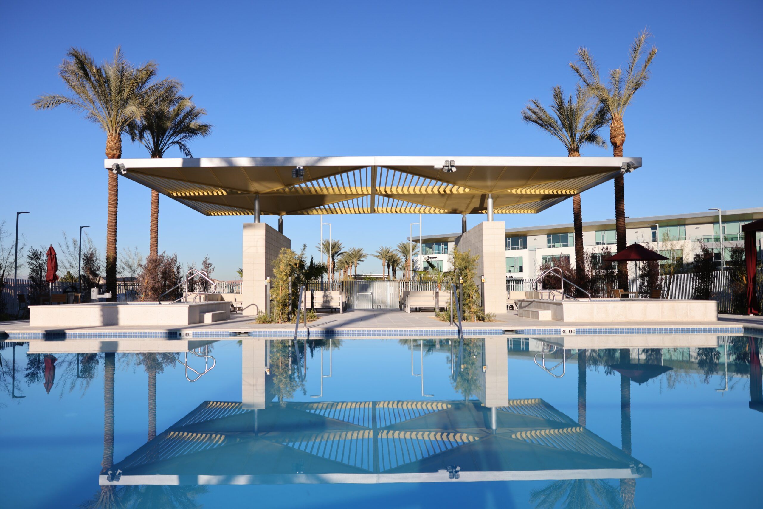A modern outdoor pool area with palm trees, cabanas, and clear blue sky reflected in the water.
