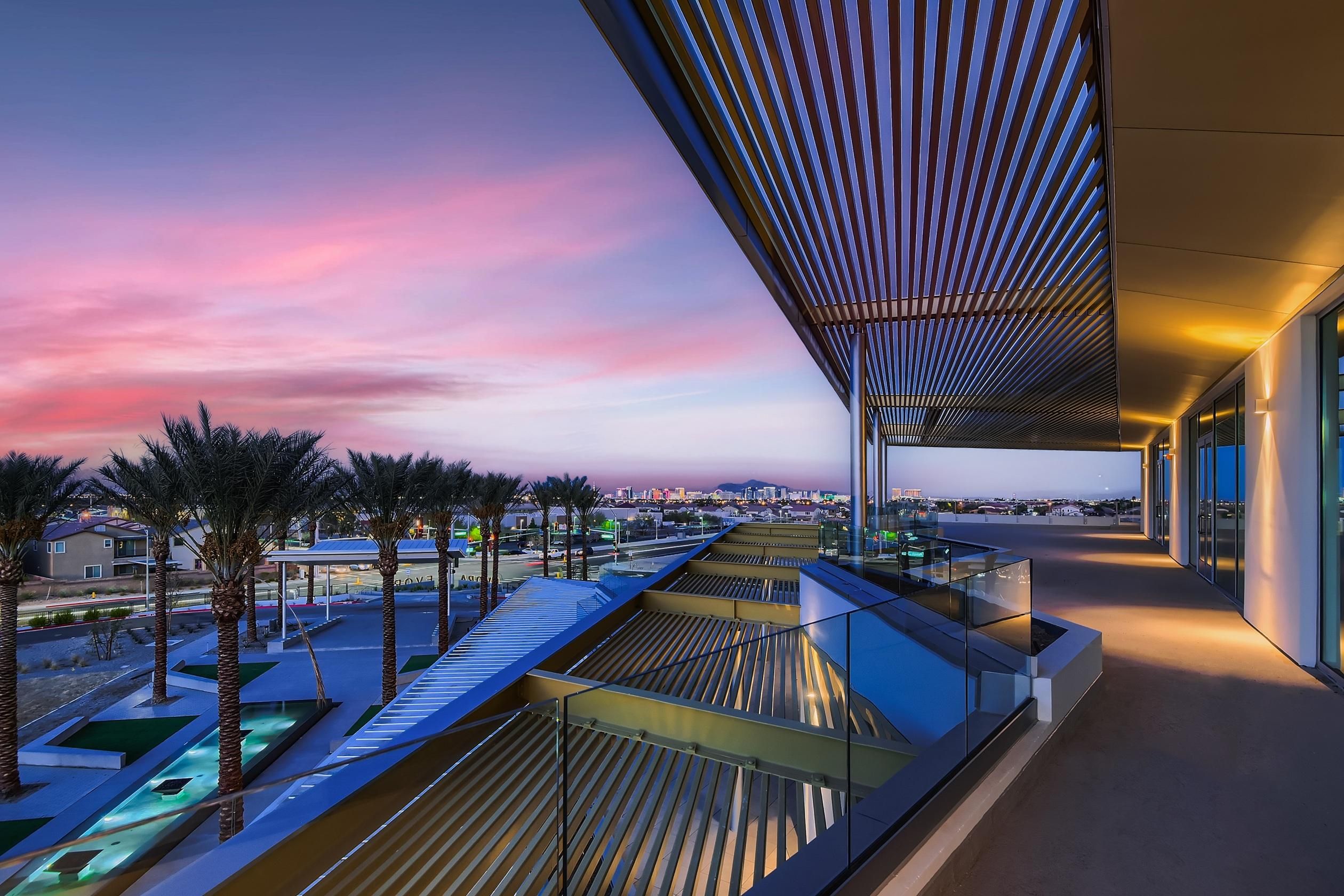 Modern balcony view at sunset with palm trees, pool area, and distant city skyline under a colorful sky.