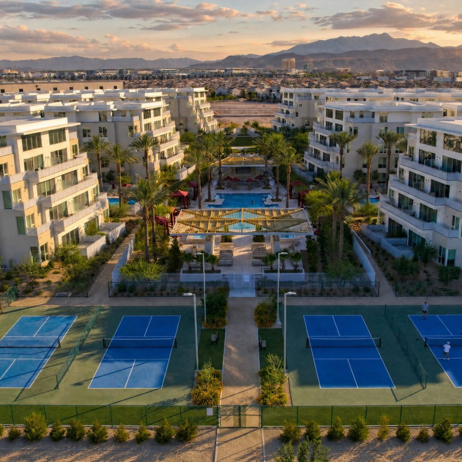 Aerial view of apartment buildings with tennis courts, a pool, and mountains in the background at sunset.